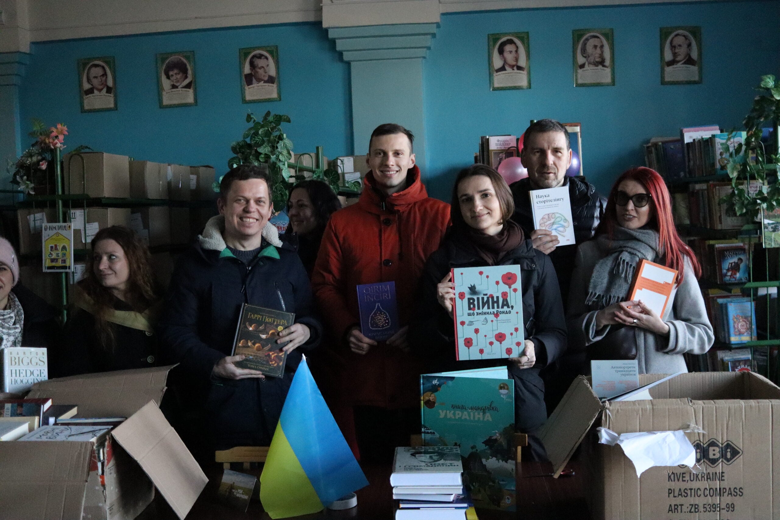 A group of people stand in a library, smiling and holding books. Boxes of books and a small Ukrainian flag are on the table in front of them. Shelves with books and portraits are in the background.