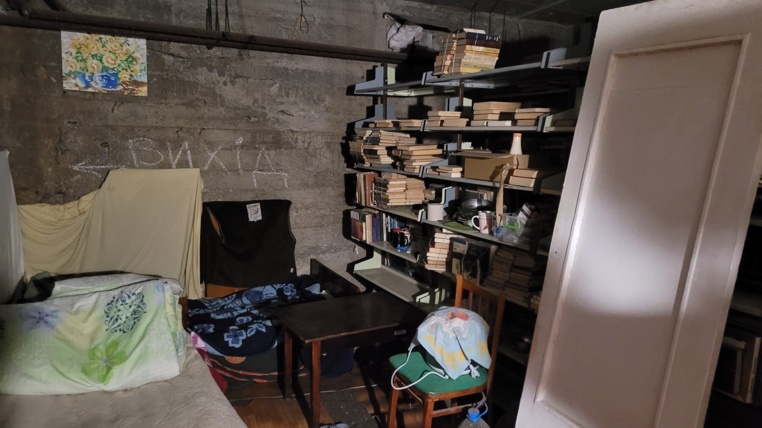 A dimly lit room with concrete walls, a bookshelf filled with books, makeshift bedding, tables and chairs, and a door leaning against the wall. Вихід (exit) with an arrow is written in chalk on the wall.