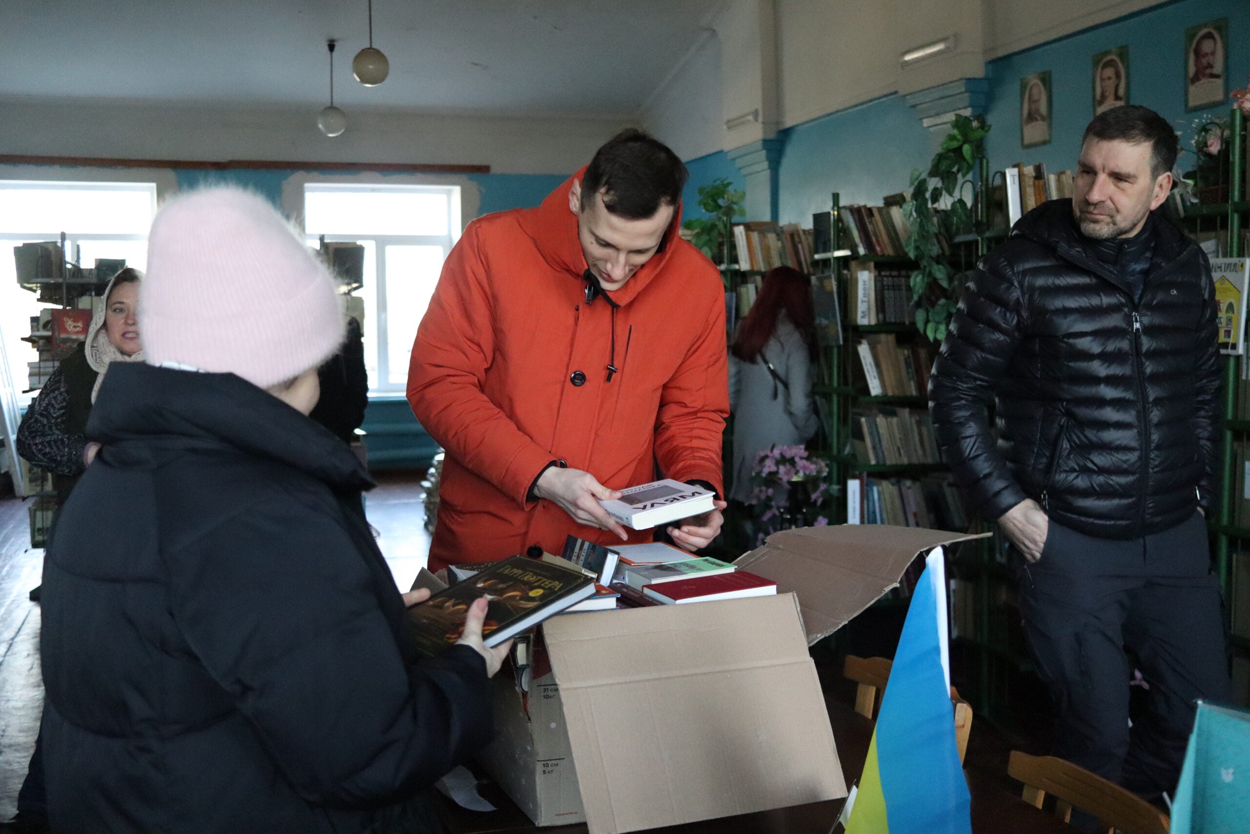 People gather around a box of books in a library. One man in a red jacket examines a book, while others look on. Bookshelves and a Ukrainian flag are visible in the background.