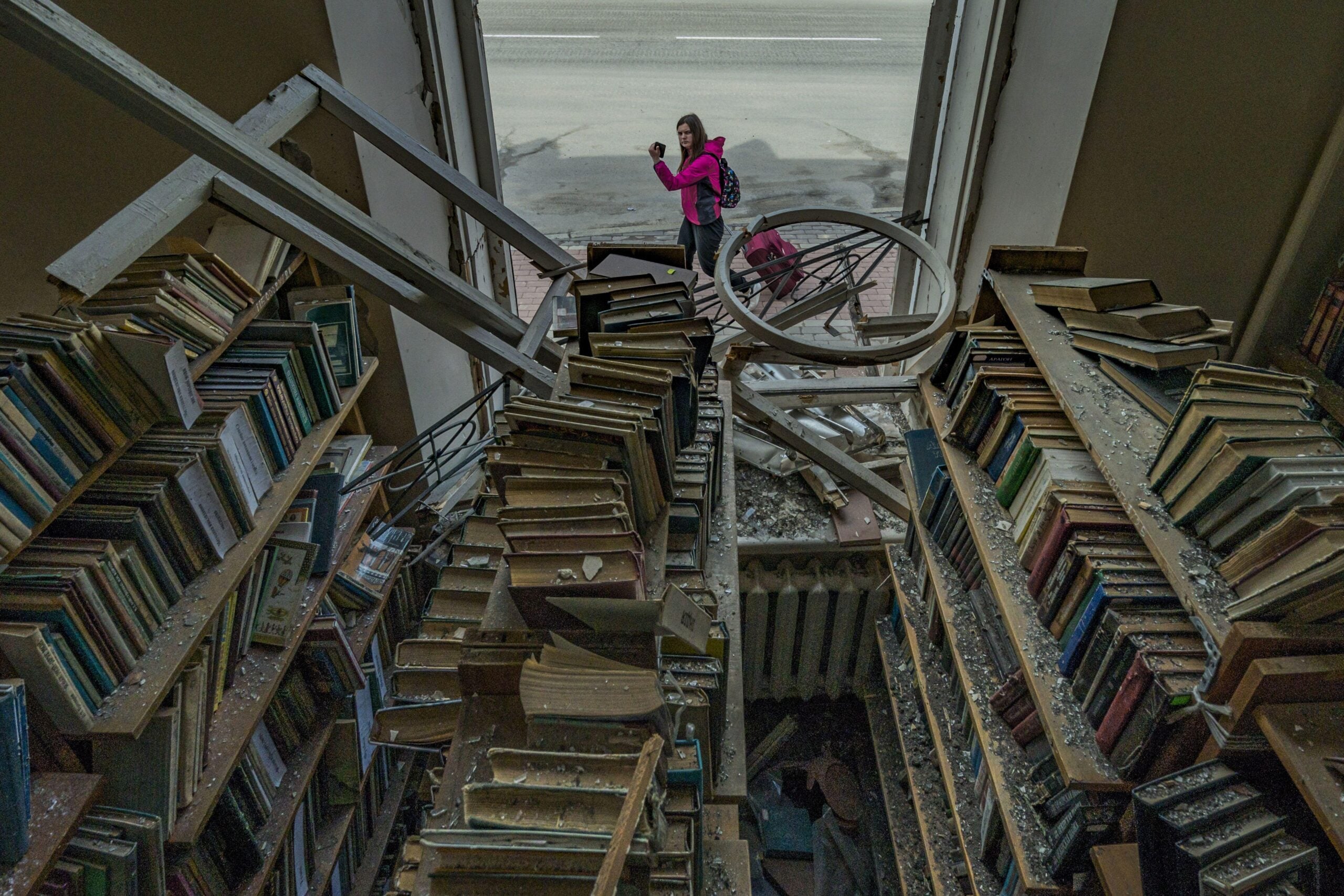 Two people stand in a damaged, dusty library filled with disorganized bookshelves. Debris and broken beams litter the area, while light enters from a large doorway at the back of the scene.