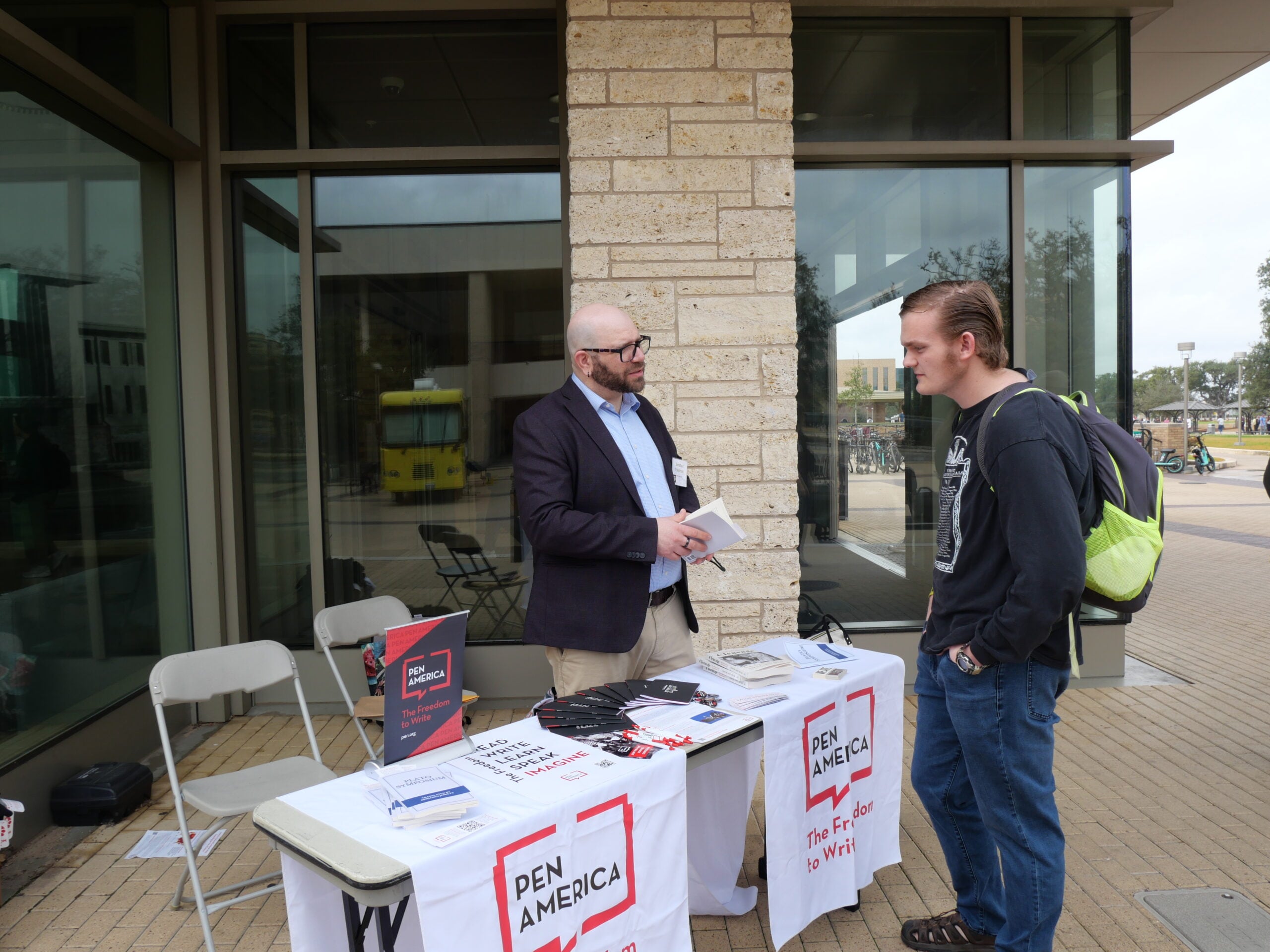 A man stands at an outdoor table covered with PEN America materials, talking to another man carrying a backpack. The table is set up in front of a building with large windows.