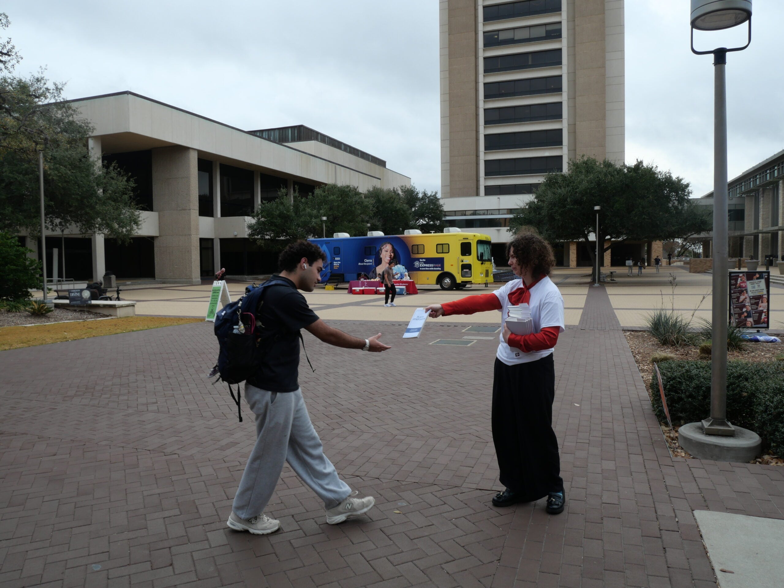 A person wearing a backpack receives a flyer from another person in a white and red shirt on a university campus walkway, with buildings, trees, and a colorful bus in the background.