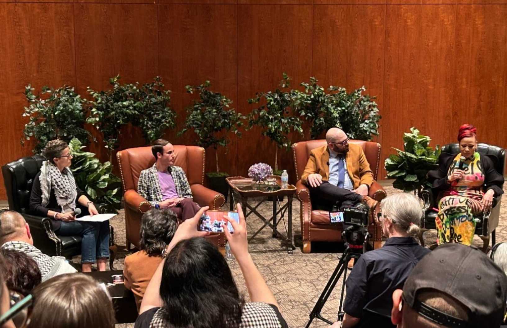 Four people sit in large chairs on a stage in front of plants and a wooden wall, having a panel discussion. Audience members watch, some filming with phones. The atmosphere is formal and attentive.