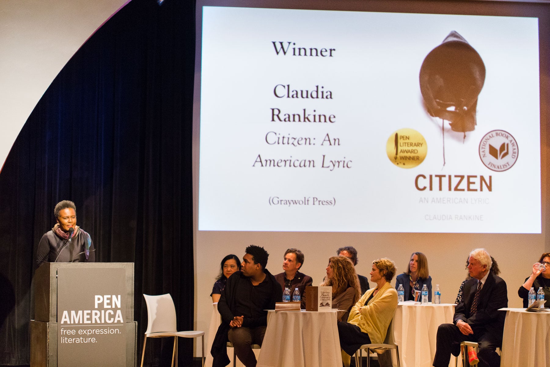 Claudia Rankine speaks at a podium labeled PEN America as she wins a literary award for Citizen: An American Lyric. Behind her, people sit at tables and the book cover is displayed on a large screen.