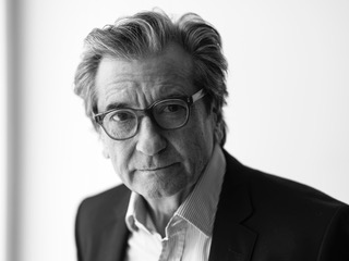 Black and white portrait of an older man with gray hair and glasses, wearing a suit jacket and collared shirt, looking thoughtfully toward the camera with a neutral background.