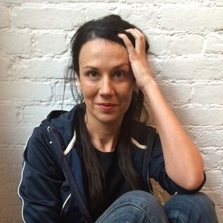 A woman with dark hair wearing a navy jacket, jeans, and red striped socks sits on a wooden floor against a white brick wall, resting her hand on her head and looking at the camera.