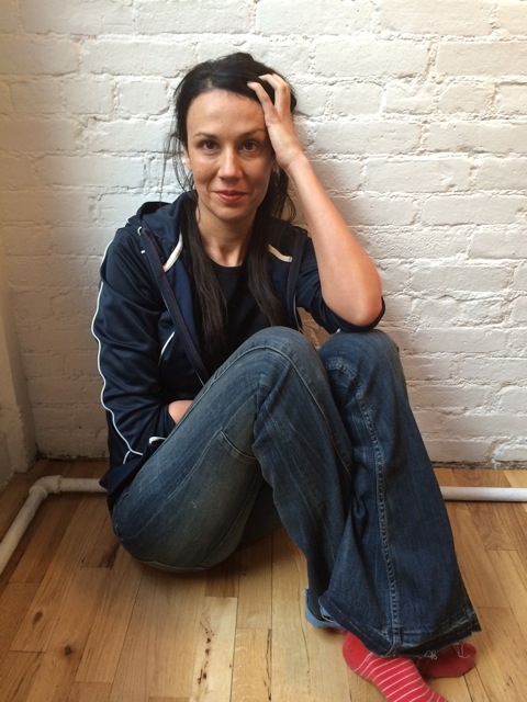 A woman with dark hair wearing a navy jacket, jeans, and red striped socks sits on a wooden floor against a white brick wall, resting her hand on her head and looking at the camera.