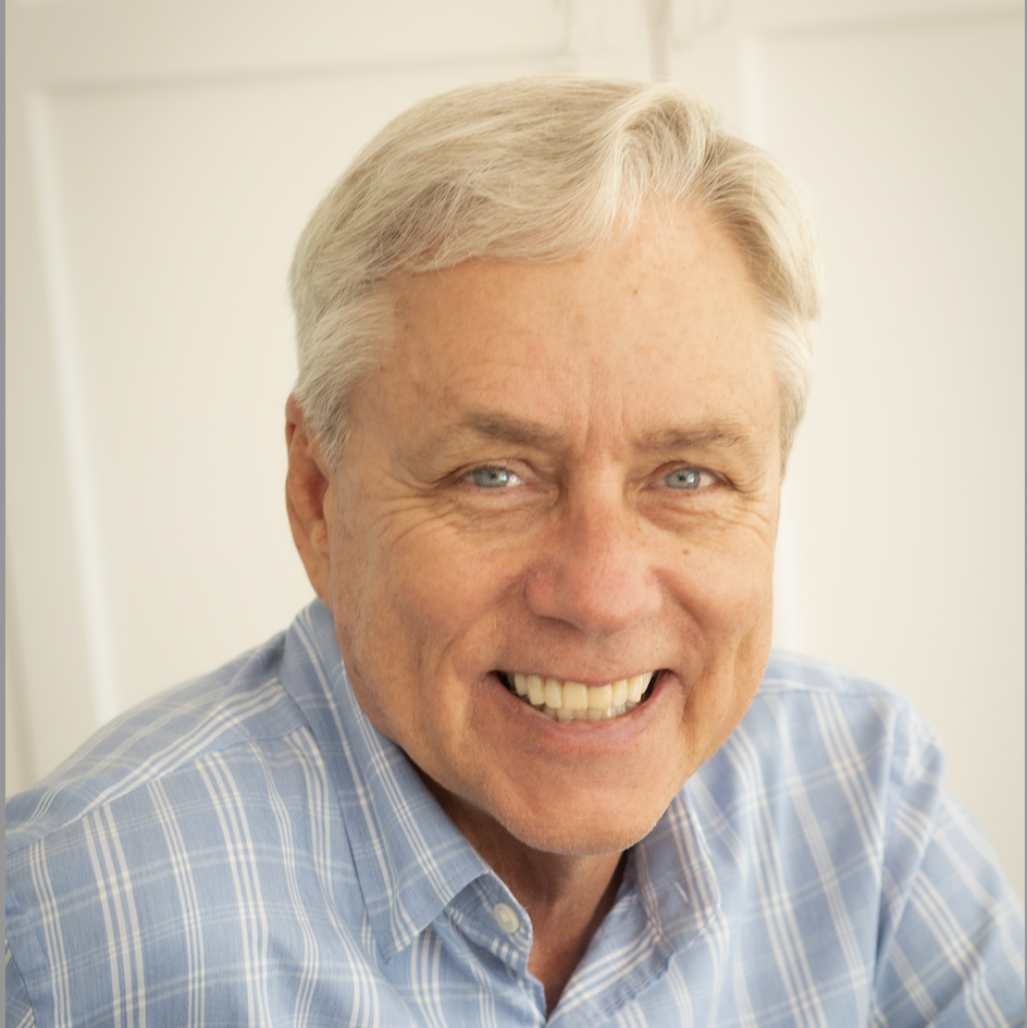 Smiling older man with short gray hair wearing a light blue plaid shirt, sitting indoors in front of a light-colored background.