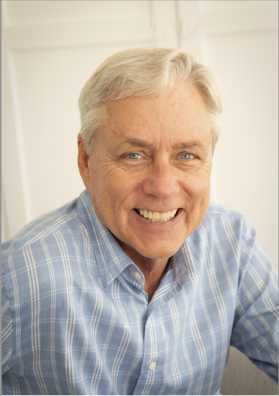 Smiling older man with short gray hair wearing a light blue plaid shirt, sitting indoors in front of a light-colored background.