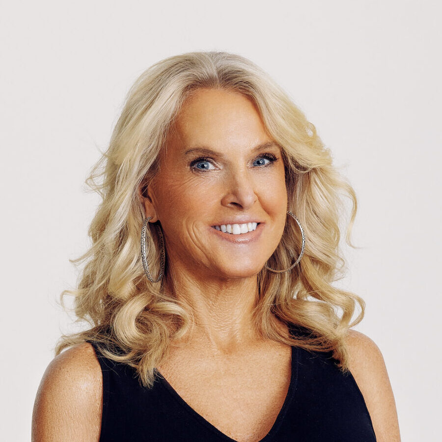 A woman with long blonde hair wearing a sleeveless black dress stands smiling with her hands clasped in front of her against a plain white background.