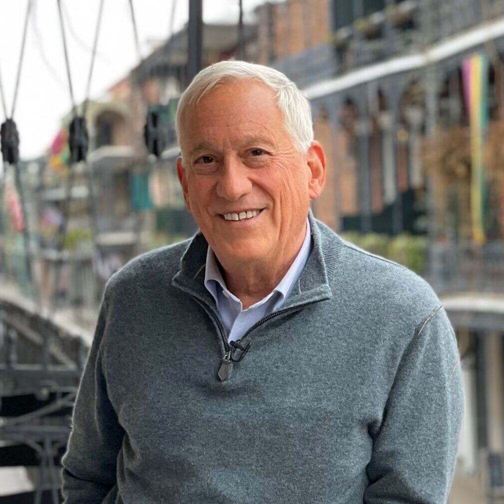 A smiling older man in a gray sweater stands on a balcony with potted plants, overlooking a street with iron railings and brick buildings, possibly in a historic district.