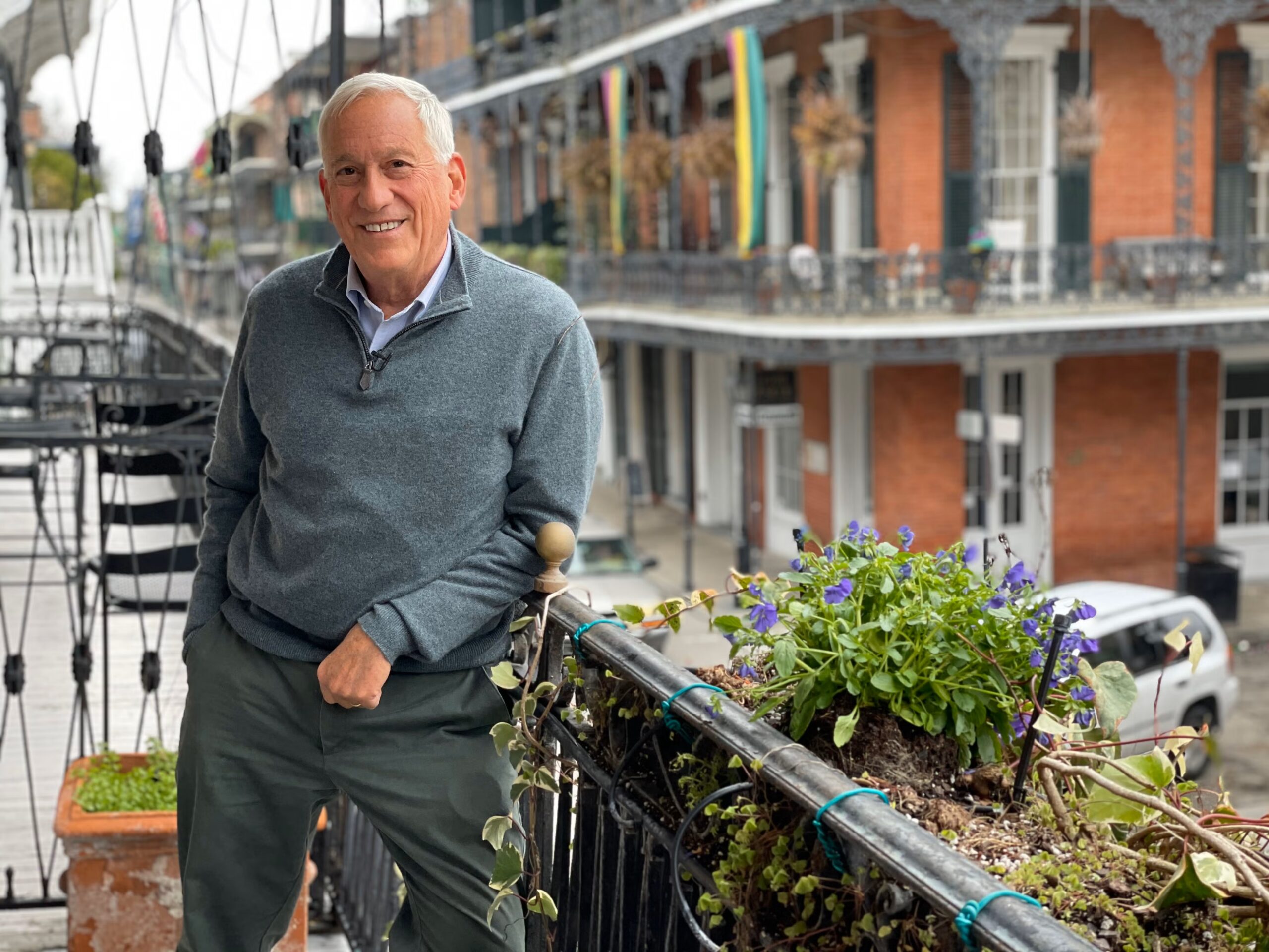 A smiling older man in a gray sweater stands on a balcony with potted plants, overlooking a street with iron railings and brick buildings, possibly in a historic district.