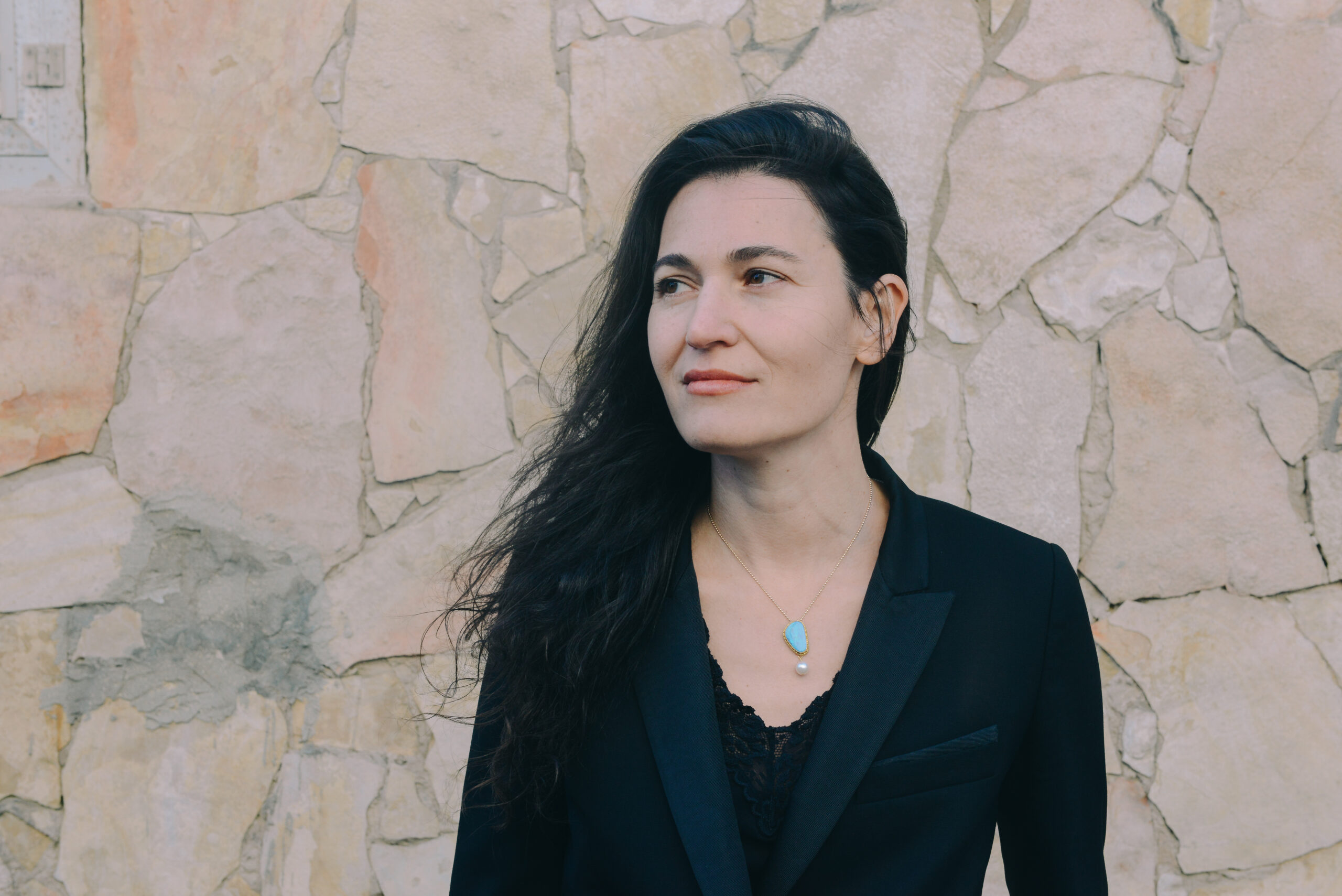 A woman with long dark hair wearing a black blazer and a turquoise pendant stands in front of a stone wall, looking slightly to the side with a neutral expression.