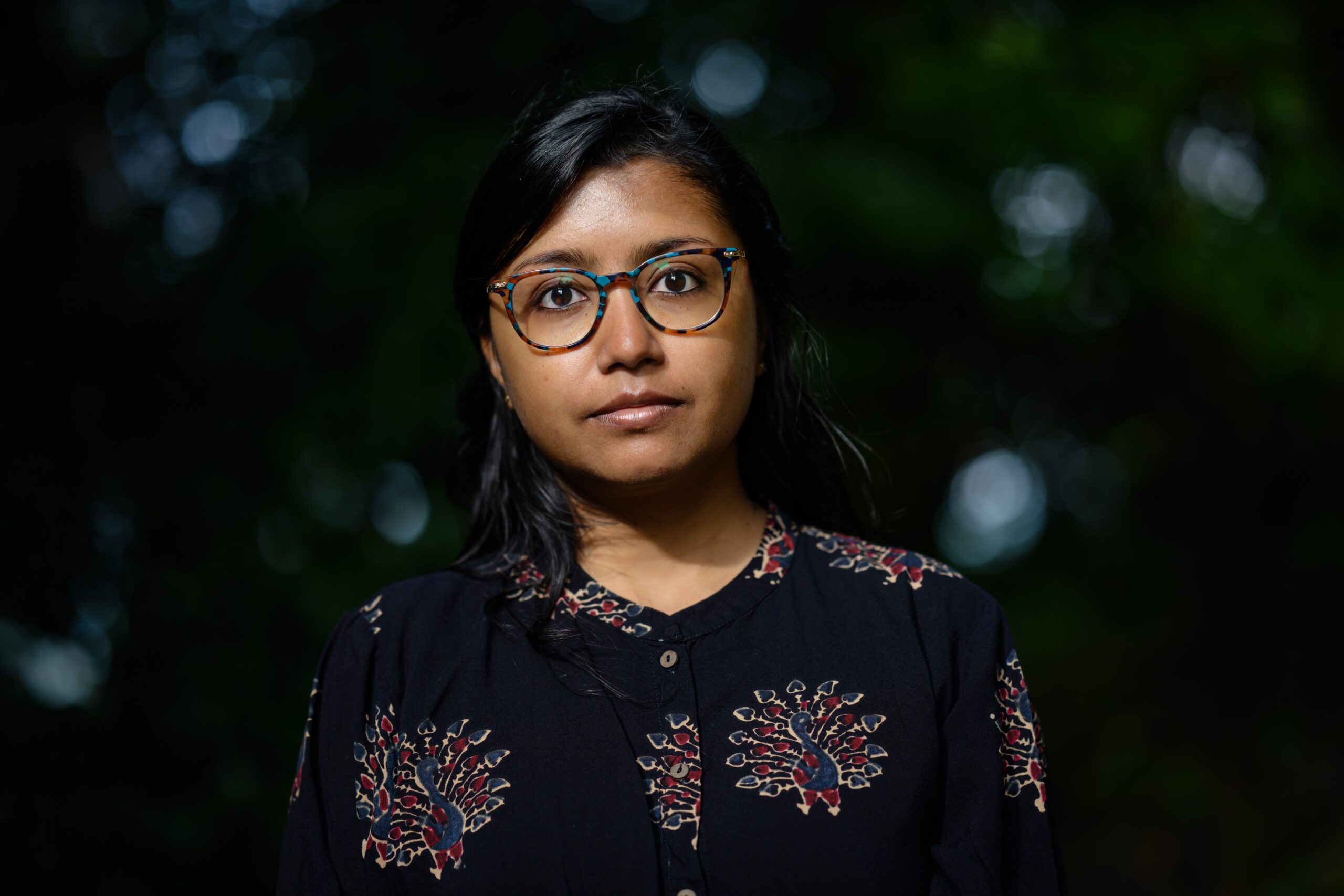 A woman with long dark hair and glasses stands outdoors against a blurred green background, wearing a black shirt with a colorful floral pattern, looking calmly at the camera.