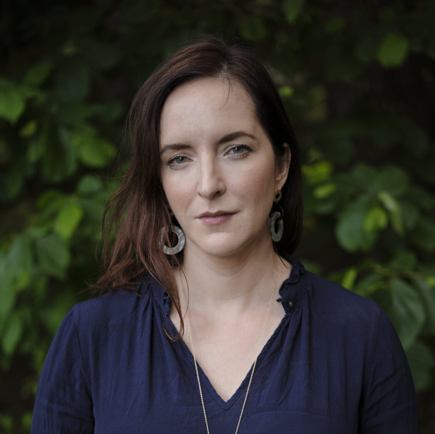 A woman with fair skin and brown hair stands outdoors in front of green foliage, wearing a navy blue blouse, circular earrings, and a long necklace with a round pendant. She looks calmly at the camera with her hands clasped.