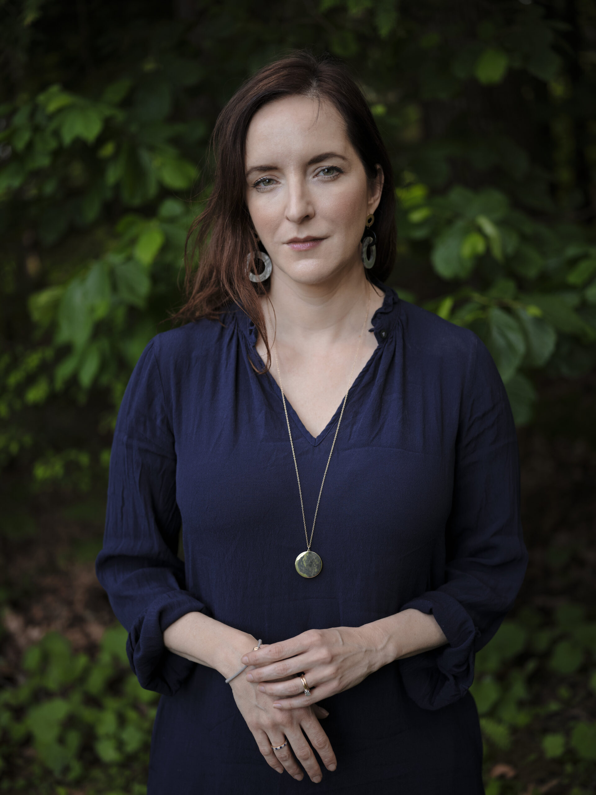 A woman with fair skin and brown hair stands outdoors in front of green foliage, wearing a navy blue blouse, circular earrings, and a long necklace with a round pendant. She looks calmly at the camera with her hands clasped.