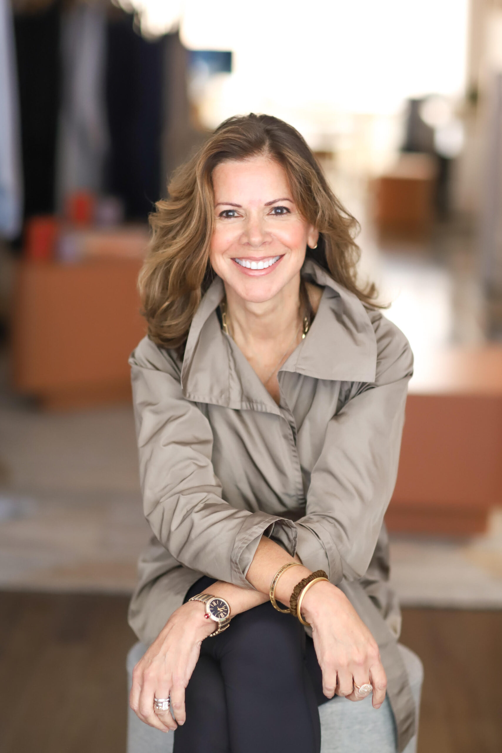 A woman with wavy brown hair, wearing a tan coat and gold jewelry, sits smiling with her arms crossed in a bright, blurred indoor setting.