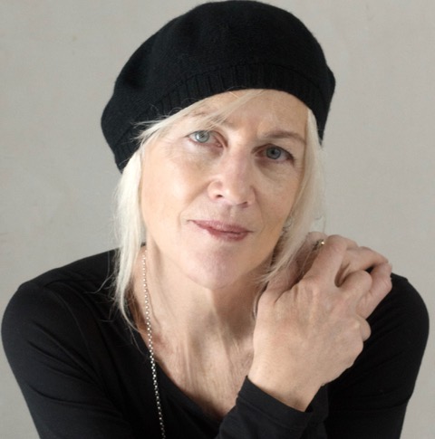 A woman with light skin and white hair wears a black beret and black top. She gazes softly at the camera, resting one hand on her shoulder against a neutral background.