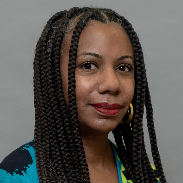A woman with long braided hair, wearing gold hoop earrings and a blue and yellow patterned top, stands against a plain gray background, looking confidently at the camera.