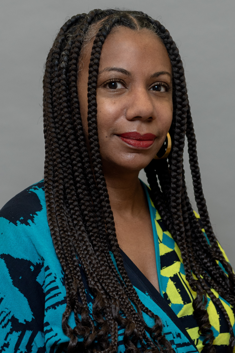 A woman with long braided hair, wearing gold hoop earrings and a blue and yellow patterned top, stands against a plain gray background, looking confidently at the camera.