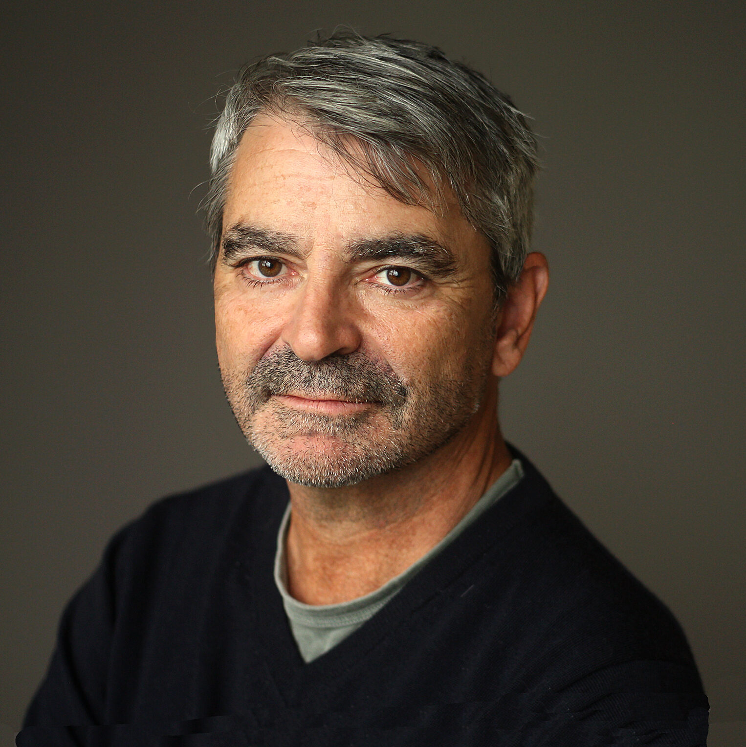 A middle-aged man with short gray hair and a salt-and-pepper beard looks at the camera with a neutral expression. He is wearing a dark sweater over a gray shirt against a plain, dark background.