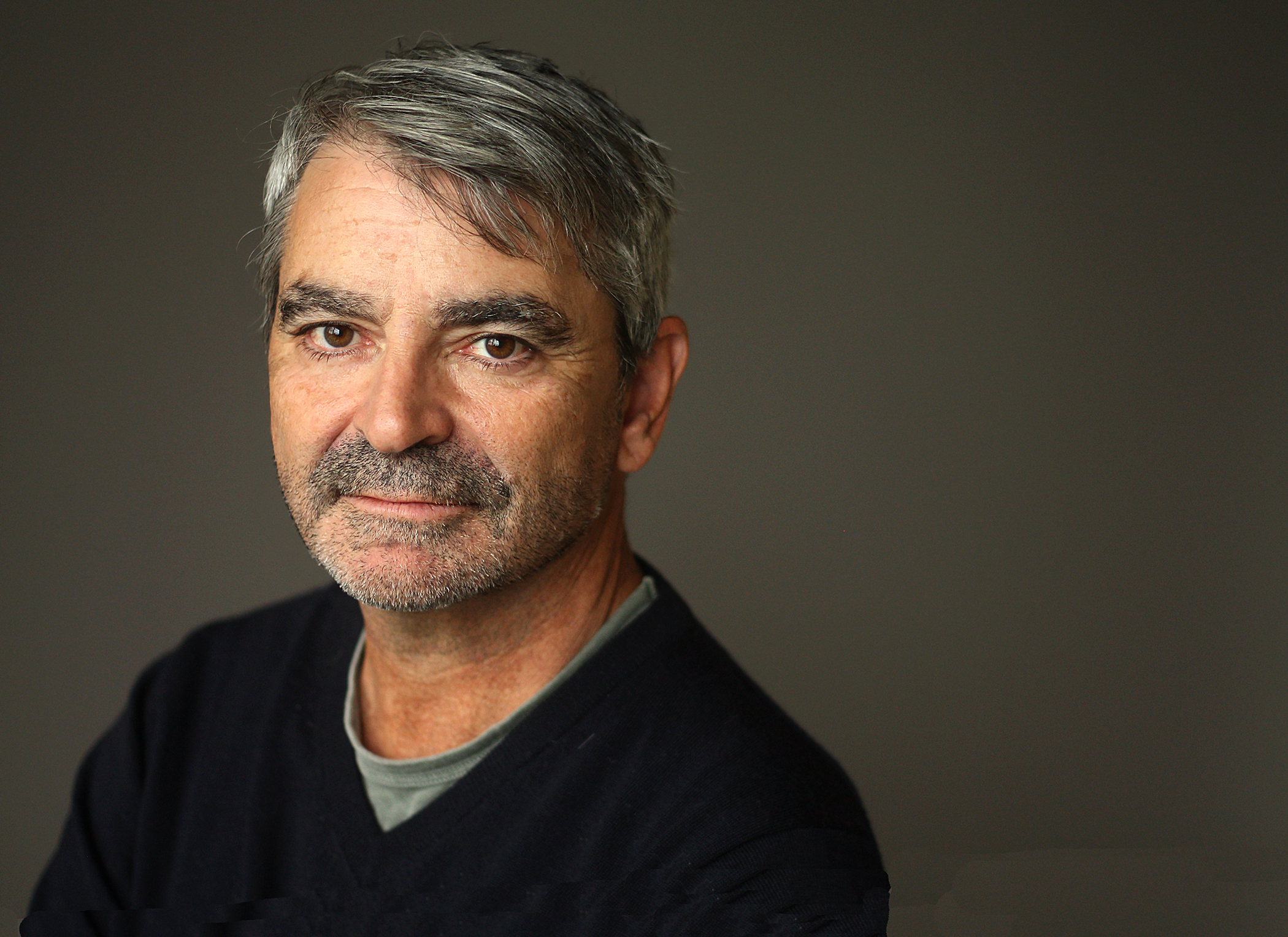 A middle-aged man with short gray hair and a salt-and-pepper beard looks at the camera with a neutral expression. He is wearing a dark sweater over a gray shirt against a plain, dark background.