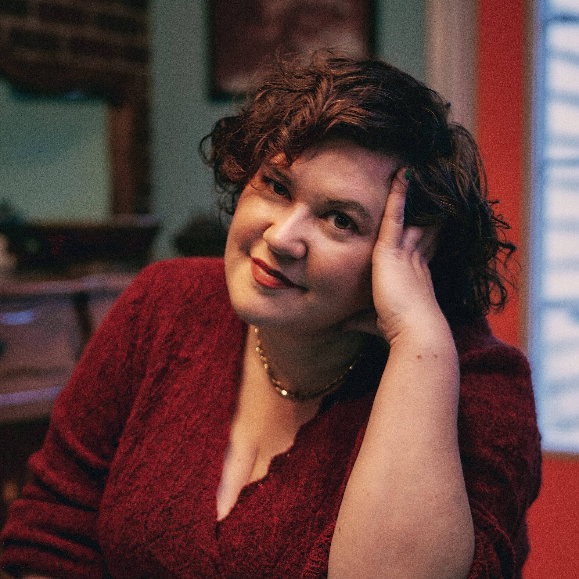 A person with short curly brown hair, wearing a red sweater, sits at a blue table indoors, resting their head on one hand and smiling softly at the camera. Warm and colorful room background.