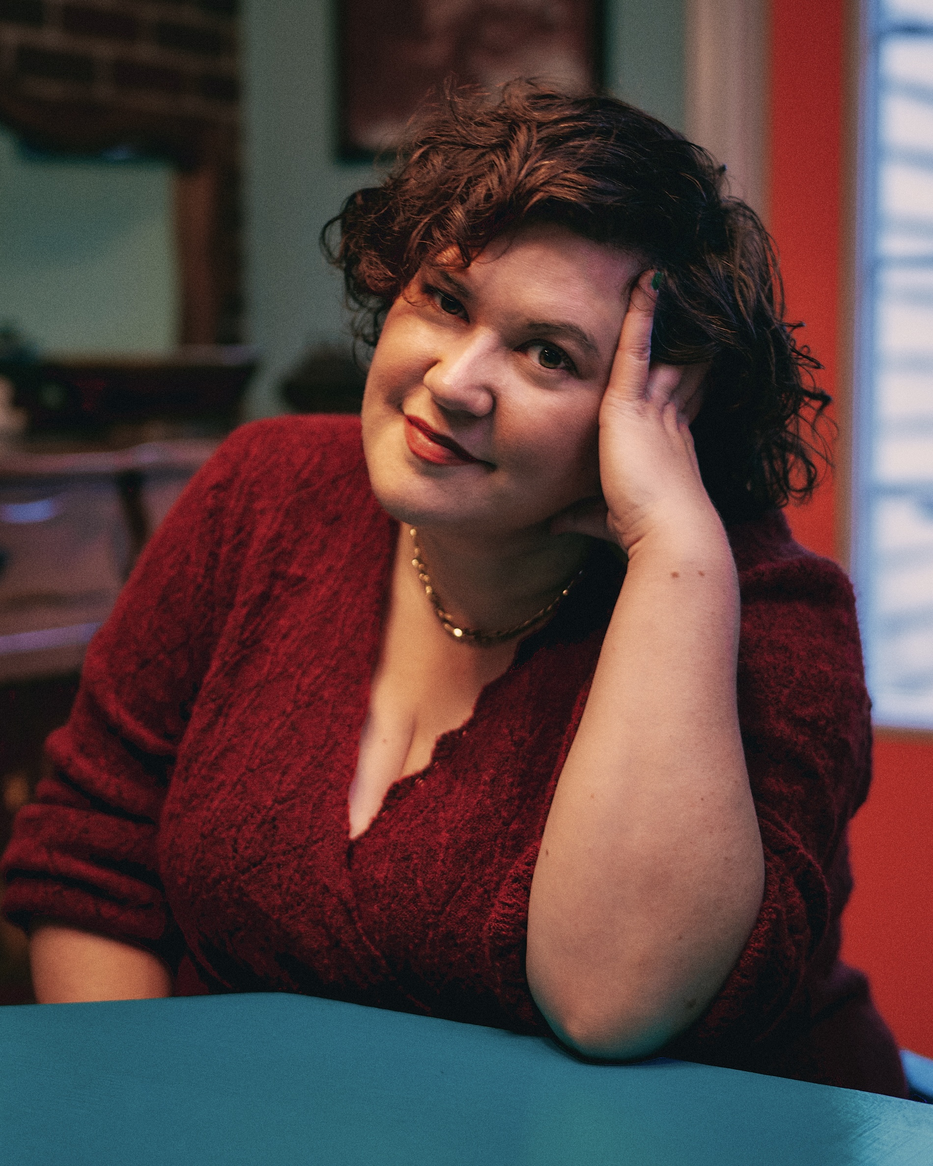 A person with short curly brown hair, wearing a red sweater, sits at a blue table indoors, resting their head on one hand and smiling softly at the camera. Warm and colorful room background.