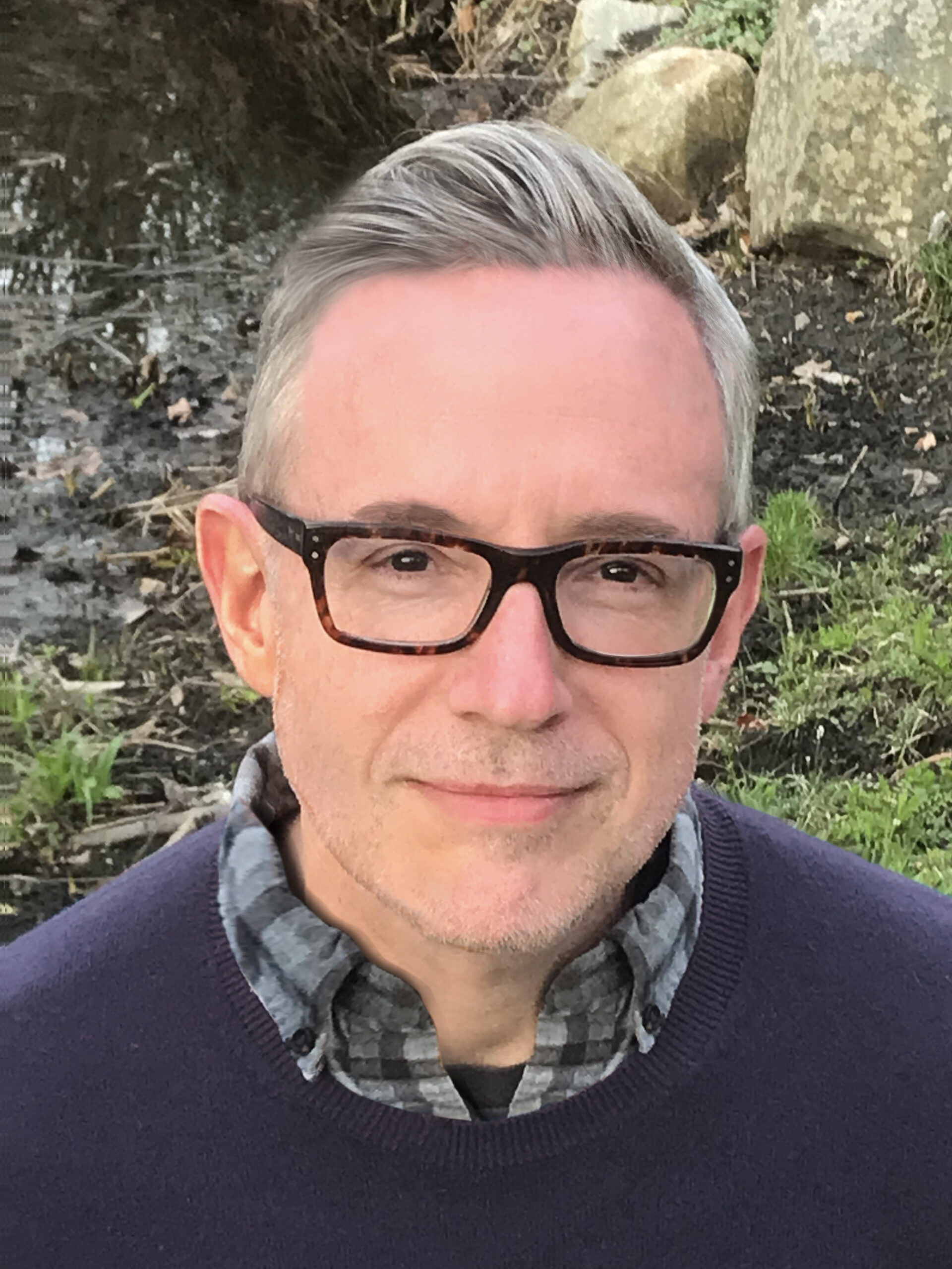 A middle-aged person with short gray hair and black glasses, wearing a plaid shirt and dark sweater, stands outdoors near grass, rocks, and a pond.