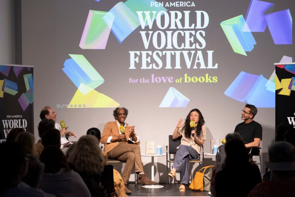 Four panelists sit on stage speaking to an audience at the PEN America World Voices Festival, with a large screen behind them displaying the festival logo and colorful book graphics.