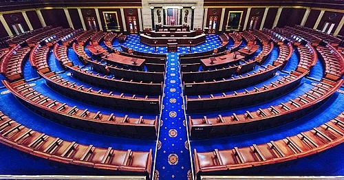 Empty legislative chamber with rows of brown desks and chairs arranged in a semicircle on a blue carpet, facing a central podium and American flag at the front.