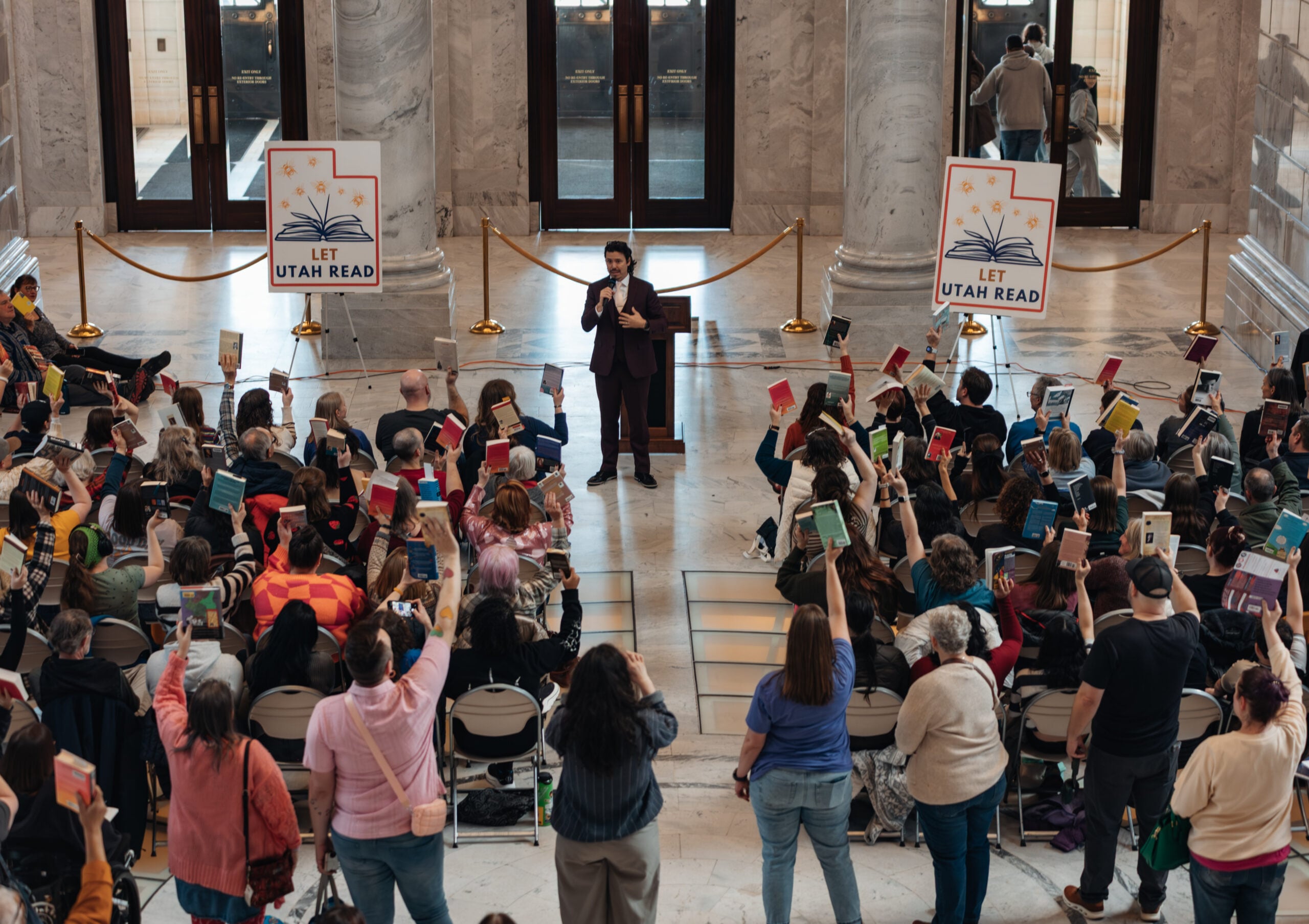 A speaker addresses a seated audience in a marble hall; many attendees hold up books. Two “Let Utah Read” signs are visible behind the speaker, and people listen attentively to the event.