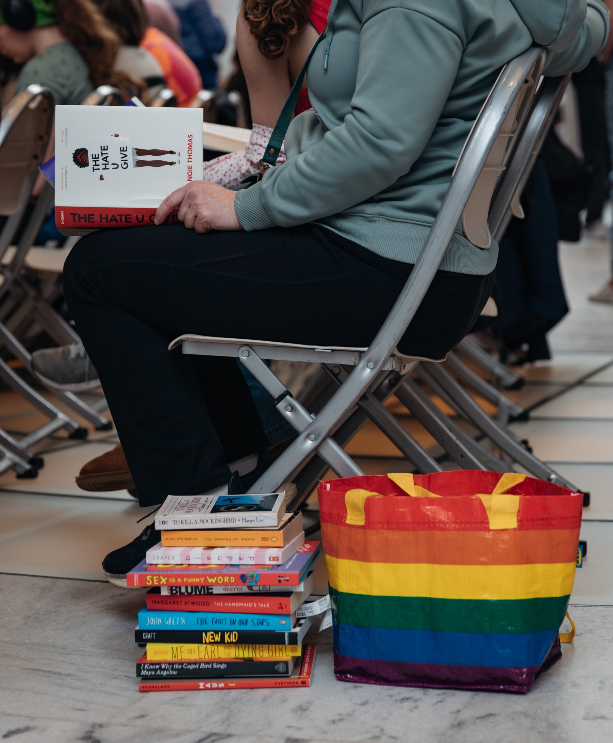 A person sits on a folding chair holding books, with a colorful rainbow tote bag and a stack of books beside them. The visible book titles include The Hate U Give and New Kid.