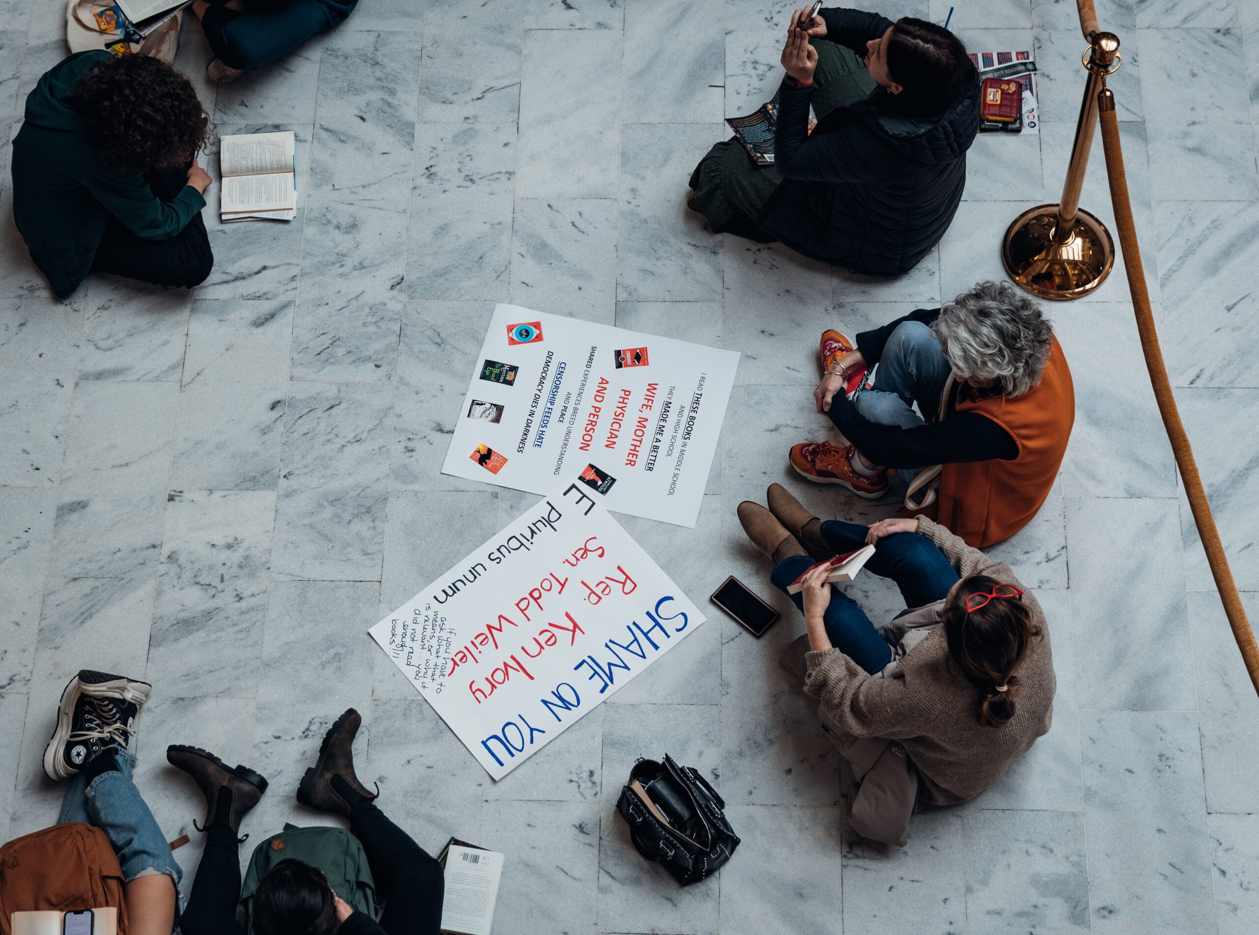 Overhead view of people sitting on a marble floor, some reading books, with protest signs in the center. One sign says SHAME ON YOU, and others mention book banning and censorship.