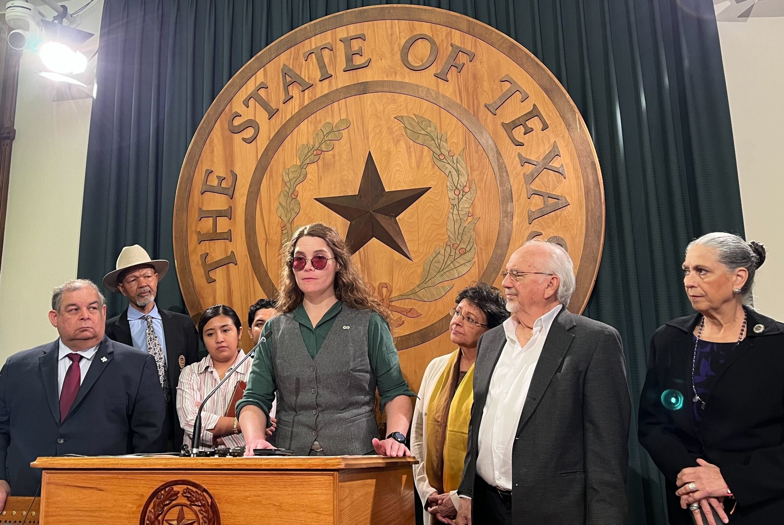 A person with long hair and glasses speaks at a podium marked with the Texas seal, surrounded by seven people. A large THE STATE OF TEXAS emblem is displayed on the wall behind them.