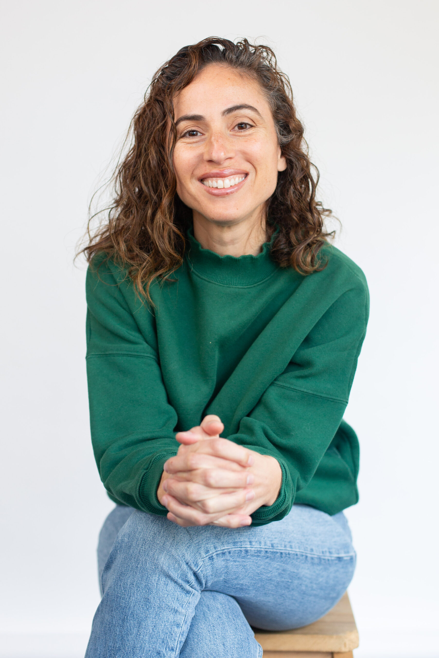 A woman with wavy brown hair wearing a green sweater and blue jeans sits on a stool, smiling at the camera with her hands clasped in front of her against a plain white background.