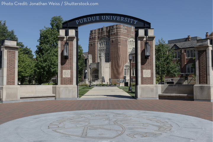 Archway entrance to Purdue University with PURDUE UNIVERSITY written on top. The brick path leads to a large building in the background, surrounded by trees and greenery under a clear sky.