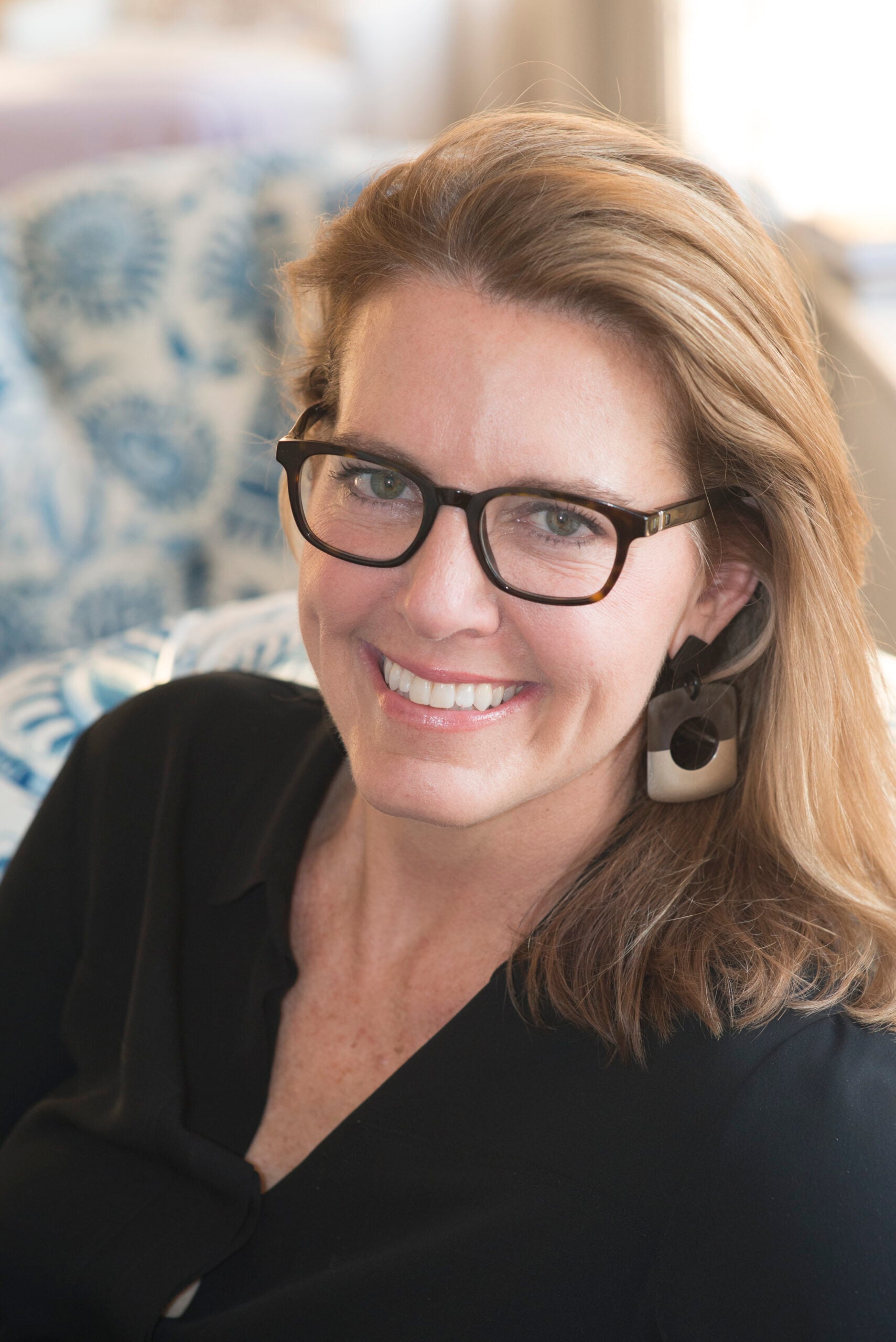 A smiling woman with light brown hair, wearing black glasses, a black top, and geometric earrings, sits indoors with a blue and white patterned background.