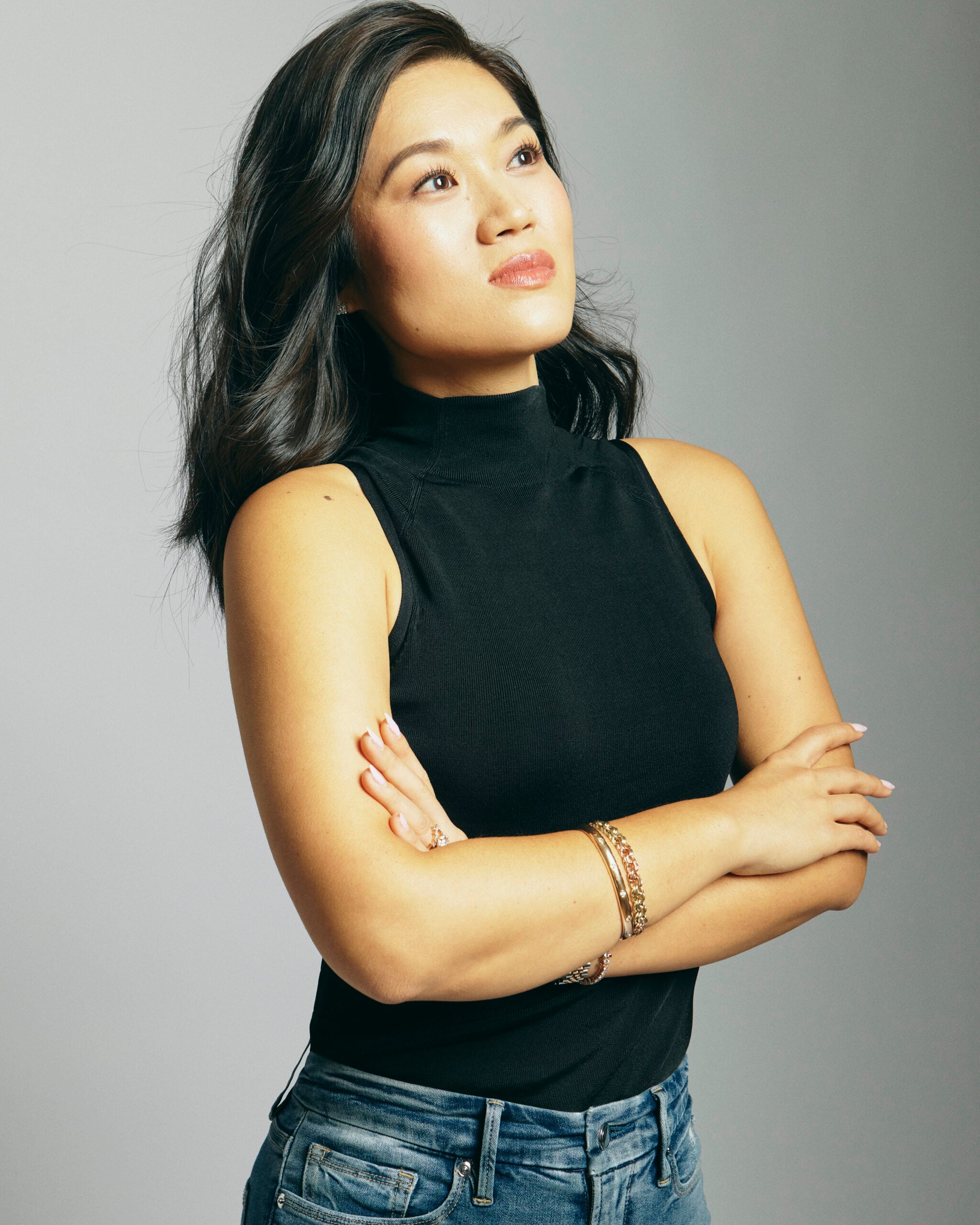 A woman with long dark hair wearing a sleeveless black top and blue jeans stands with her arms crossed, looking upward thoughtfully against a plain light gray background.
