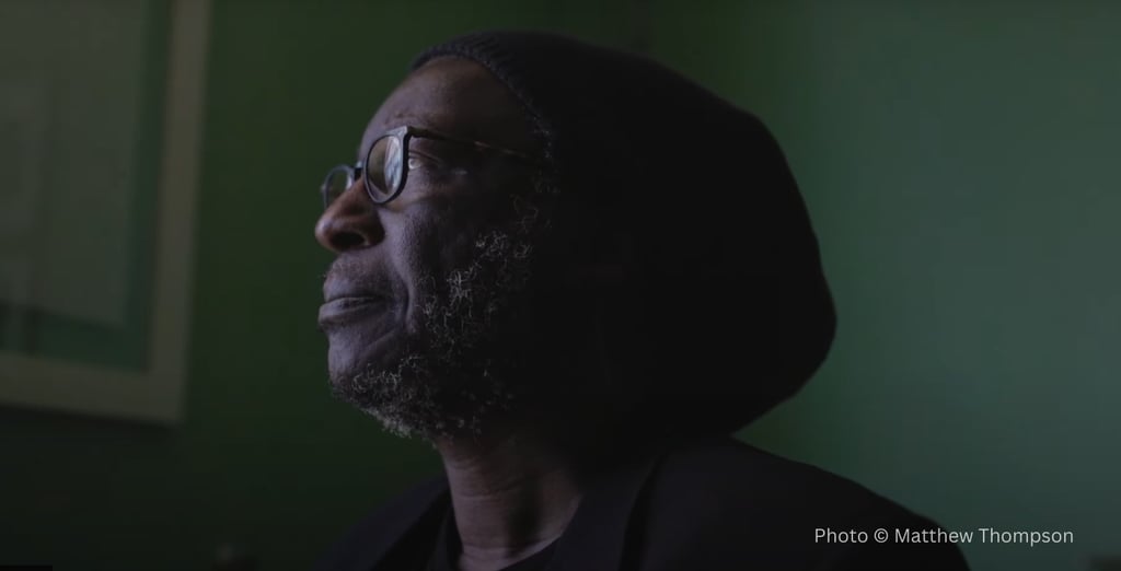 A man wearing glasses and a beanie looks thoughtfully to the left, partially illuminated by soft light against a green background. Photo © Matthew Thompson.