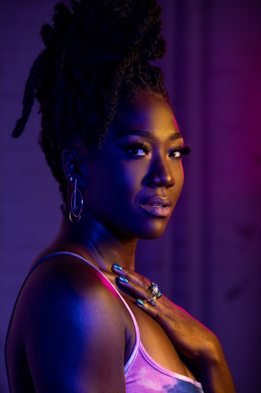 A woman with braided hair in an updo poses with her hand on her chest, wearing a tank top and hoop earrings, lit by dramatic blue and pink lighting against a dark background.