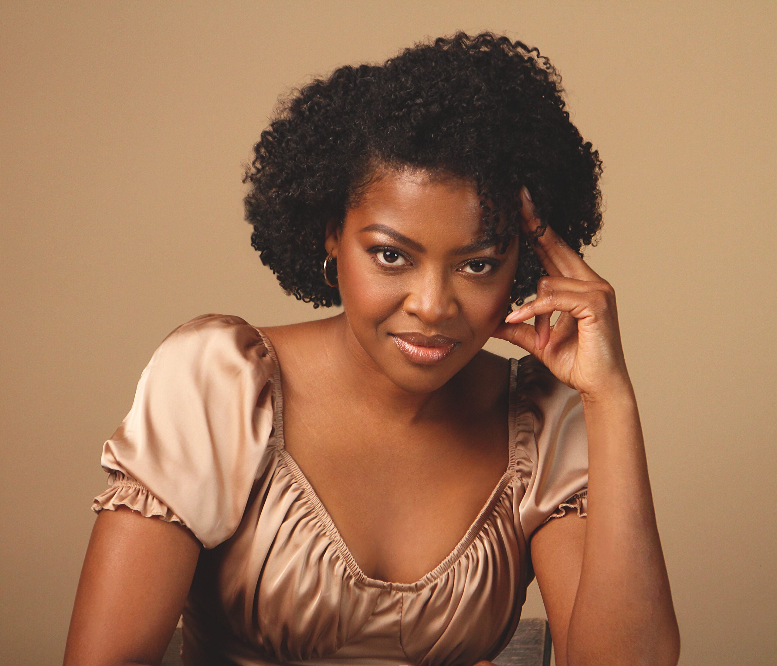 A woman with short, curly hair and hoop earrings sits against a beige background, wearing a champagne-colored, puff-sleeve blouse. She gazes confidently at the camera with one hand resting on her temple.