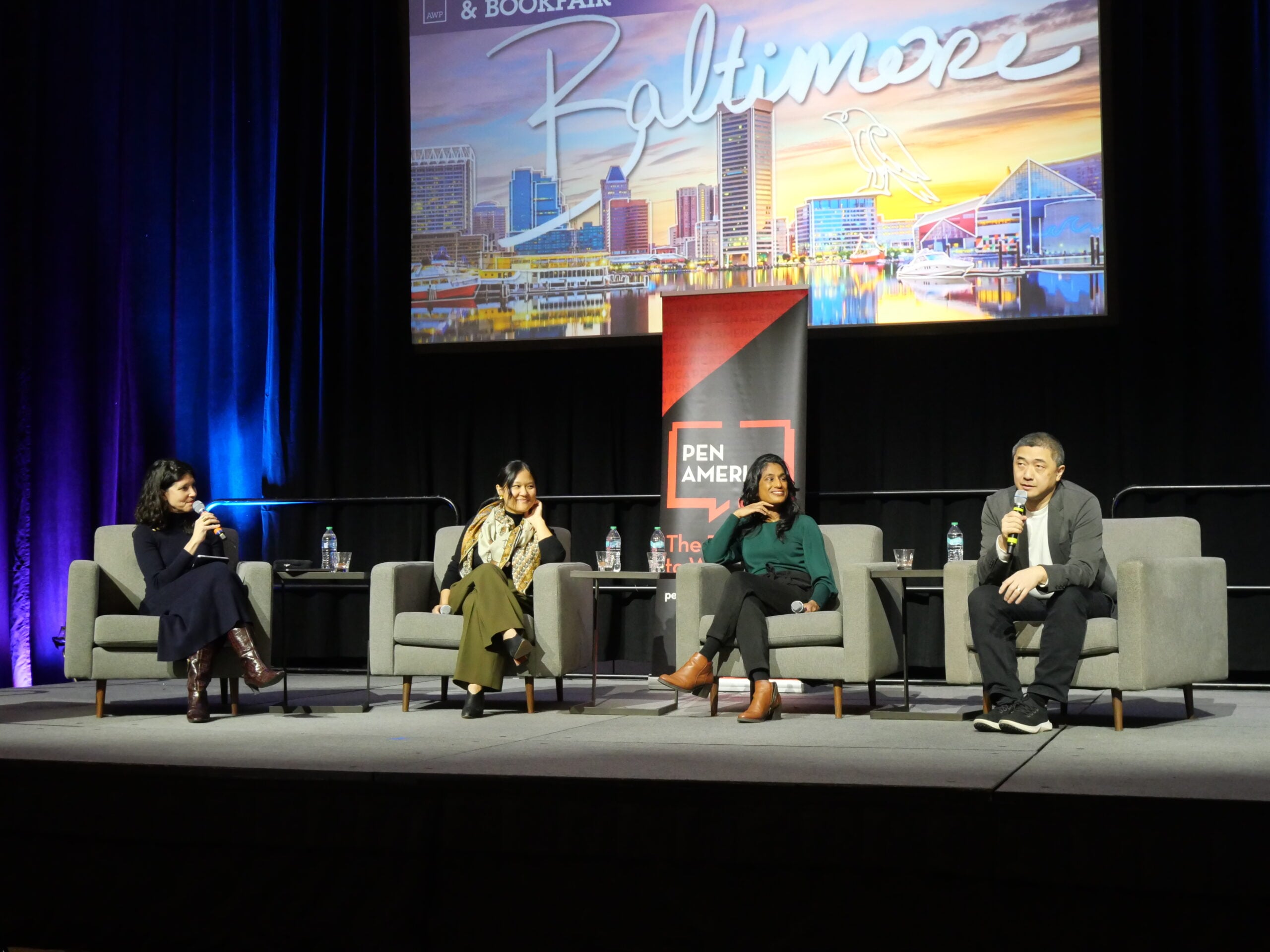 Four people sit on a stage in armchairs, holding microphones, participating in a panel discussion. A large Baltimore sign and cityscape are displayed on a screen behind them. A PEN America banner stands in the background.