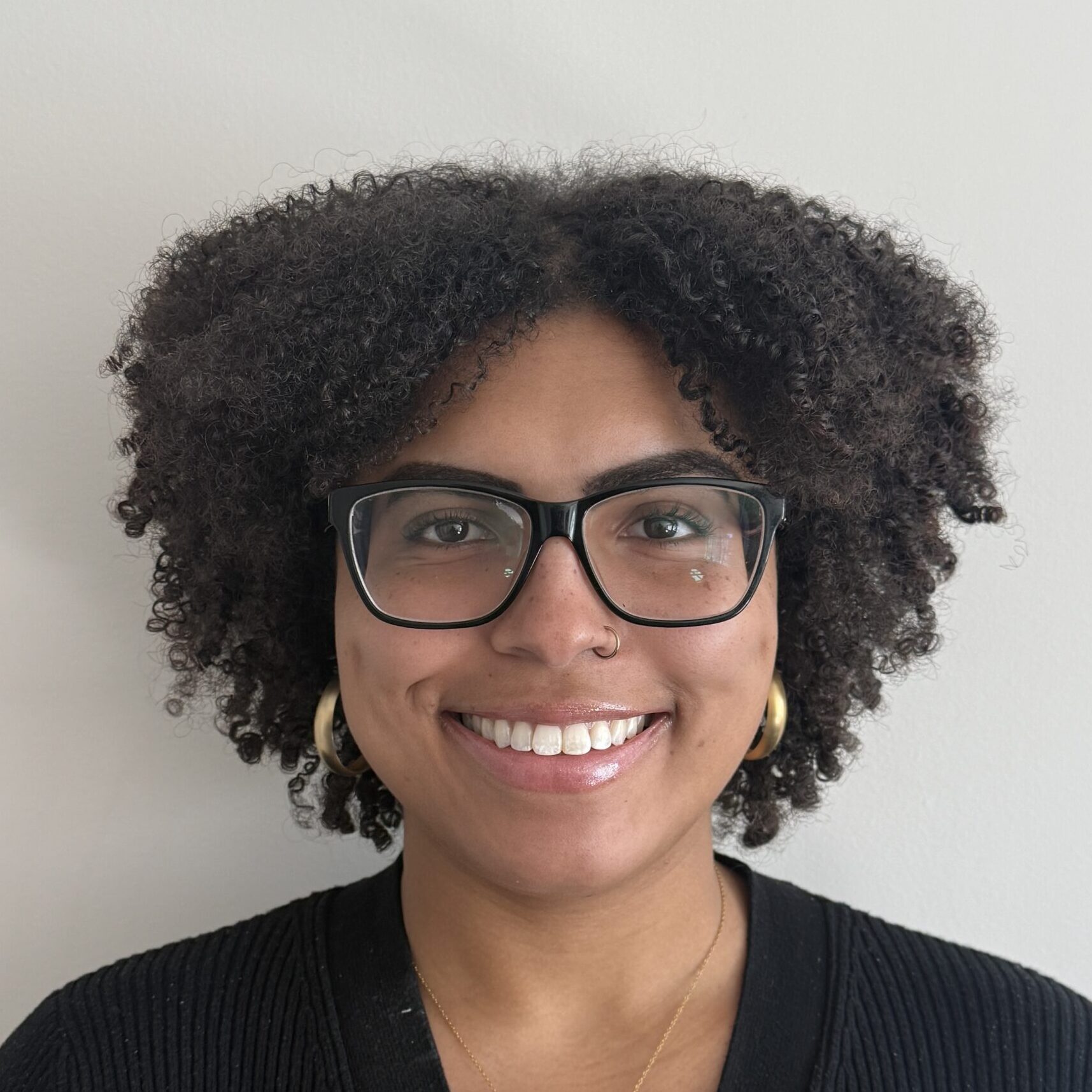 A person with curly hair, wearing glasses, gold hoop earrings, and a black top smiles at the camera in front of a plain light-colored wall.