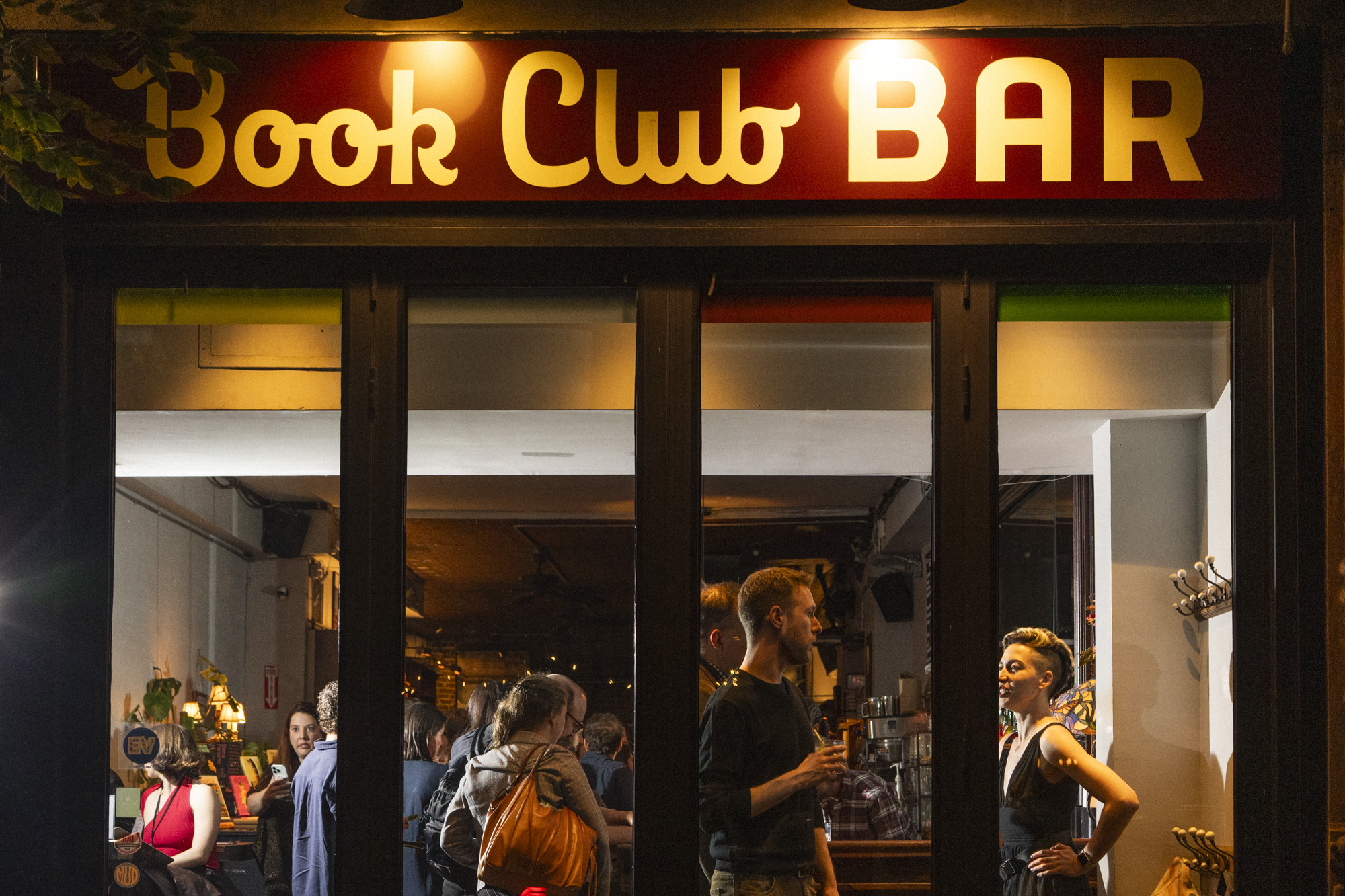 A group of people, including a young patron, socializing inside a bar with a bright red and yellow sign reading Book Club BAR above the entrance at night. Two people stand talking near the window.