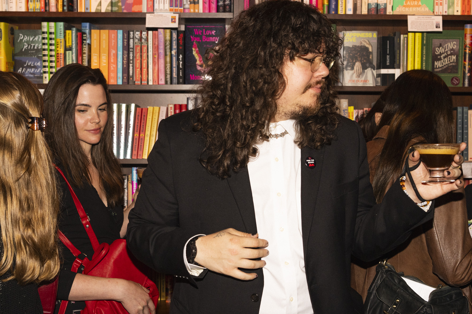 A young patron in a black suit holding a cocktail stands in front of bookshelves at a crowded bookstore event. Several people, including a woman with a red bag, are nearby, and colorful books fill the background.