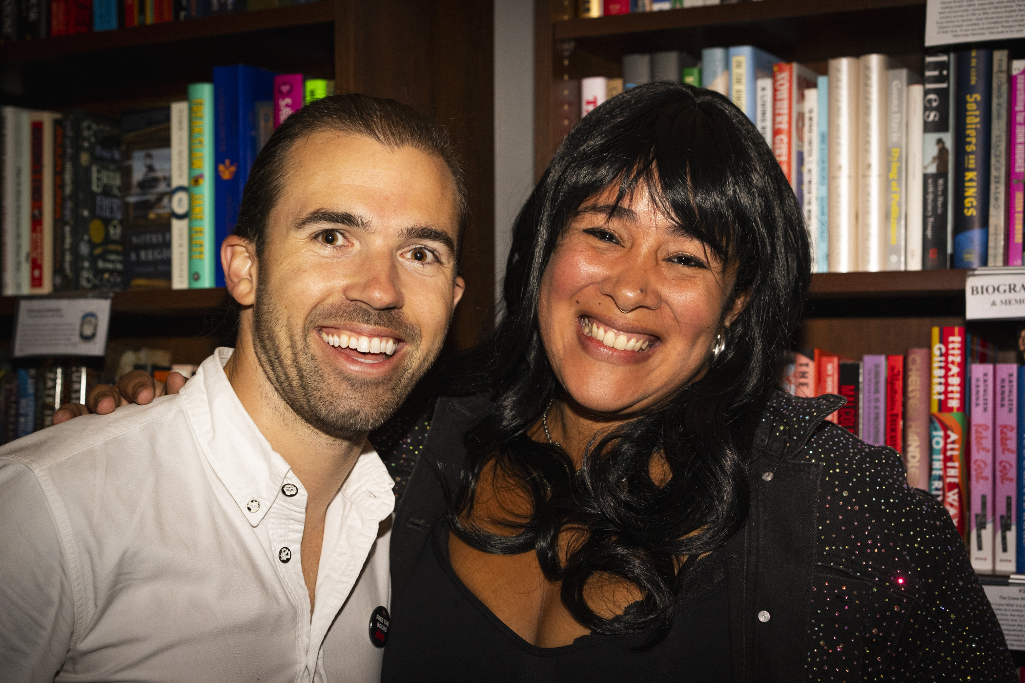 Two people, including a young patron, smile and pose together in front of bookshelves filled with colorful books. One wears a white shirt, while the other has long dark hair and a black outfit with sparkly details.