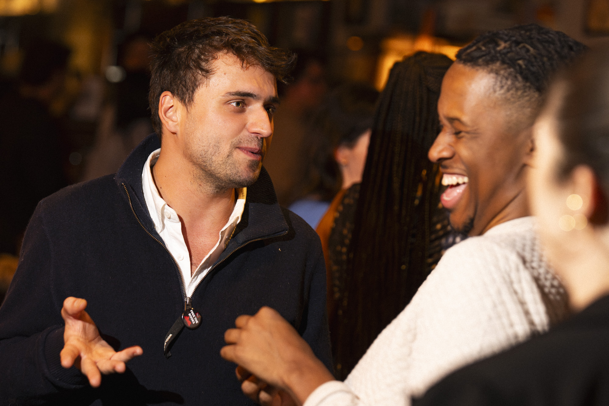Two men, including a young patron, are talking and laughing at a lively indoor social gathering. One gestures with his hand while the other smiles widely, with people and warm lighting visible in the blurred background.