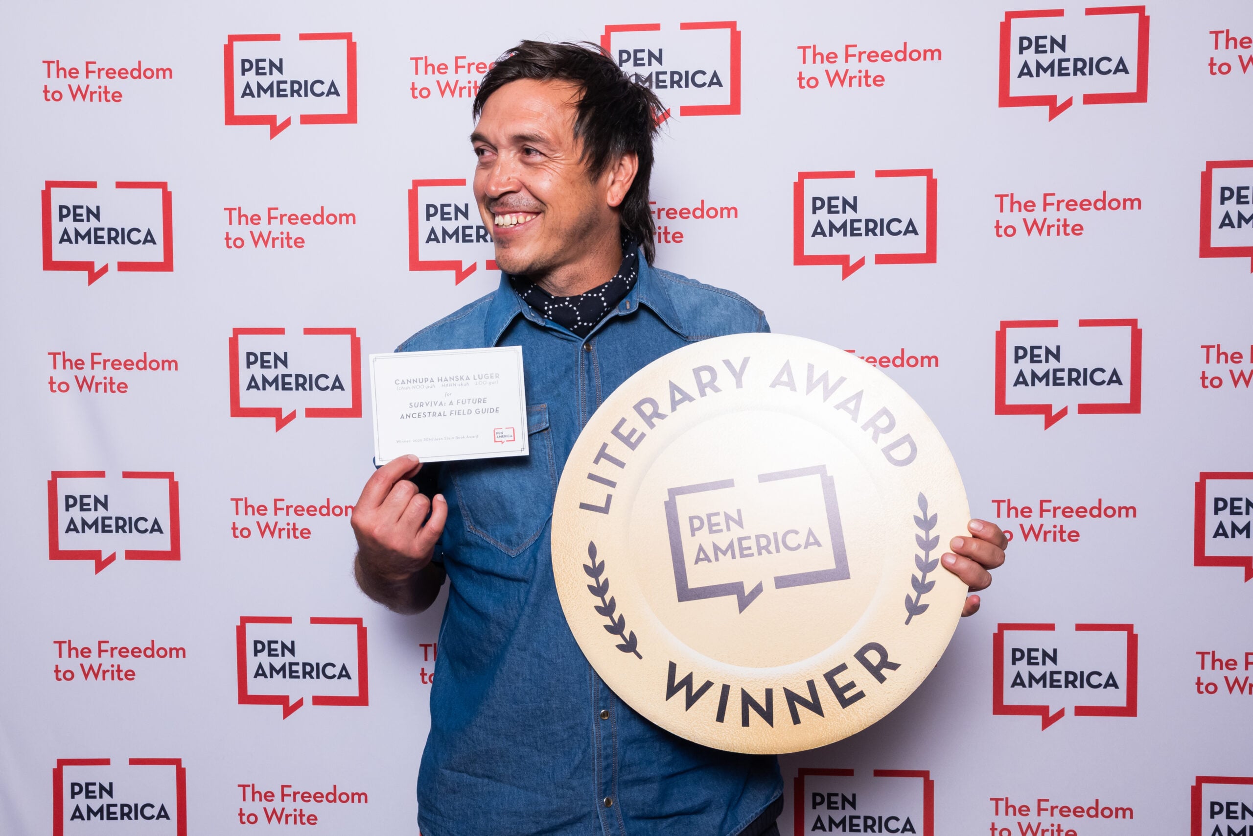 A smiling man holds a large circular “PEN America Literary Award Winner” sign and a certificate. He stands in front of a backdrop with repeated PEN America logos and the phrase The Freedom to Write.
