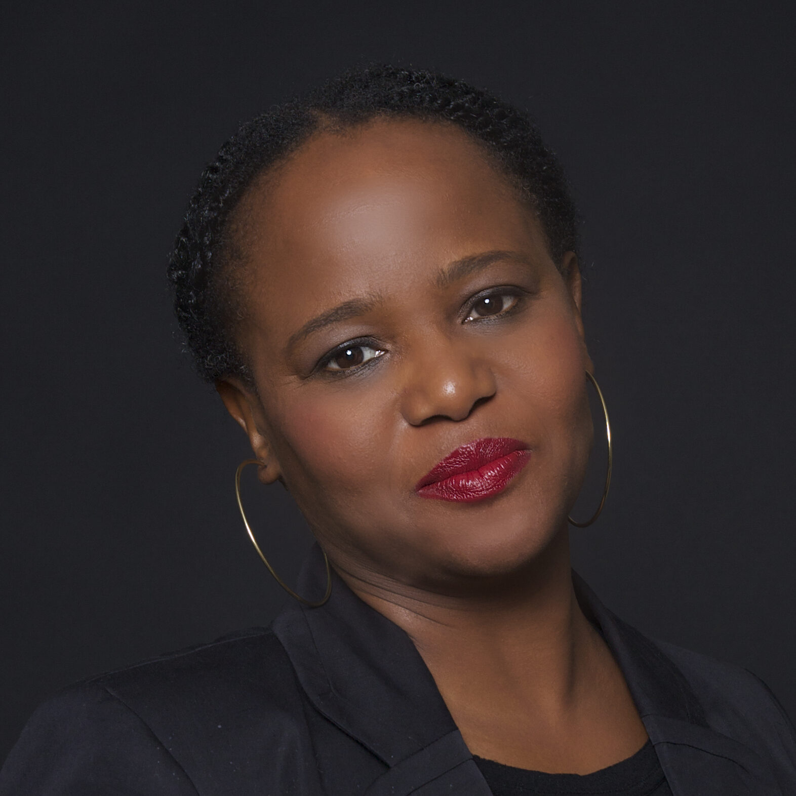 A woman with short braided hair, wearing hoop earrings, a black blazer, and a black top, poses against a dark background with a slight smile and red lipstick—her confident gaze evokes the poise of a Nabokov Award winner.